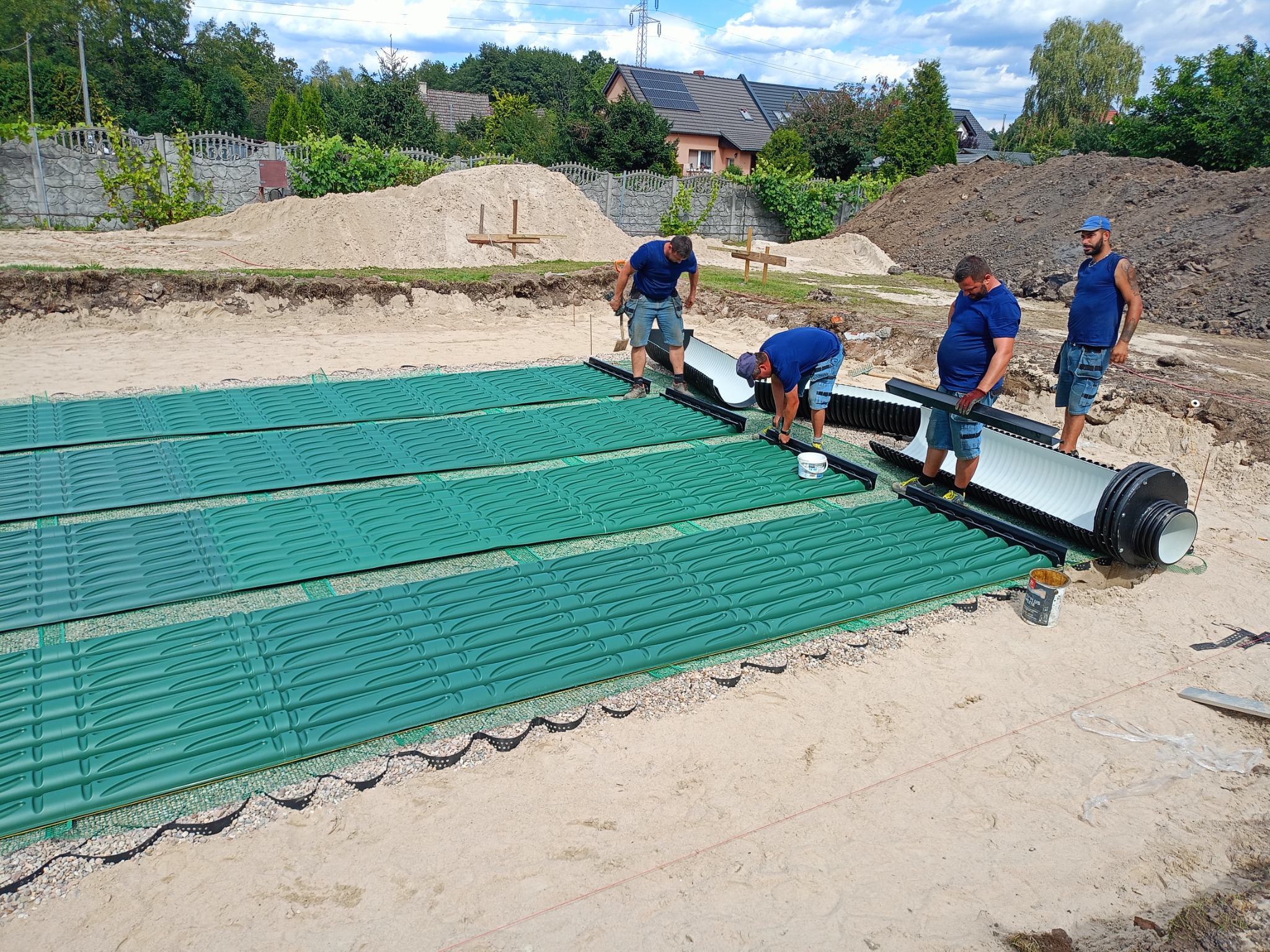 Montaż drenażu rozsączającego: czterech mężczyzn instaluje system zielonych paneli drenażowych na piaszczystym terenie budowy, widok z góry na domy w tle.