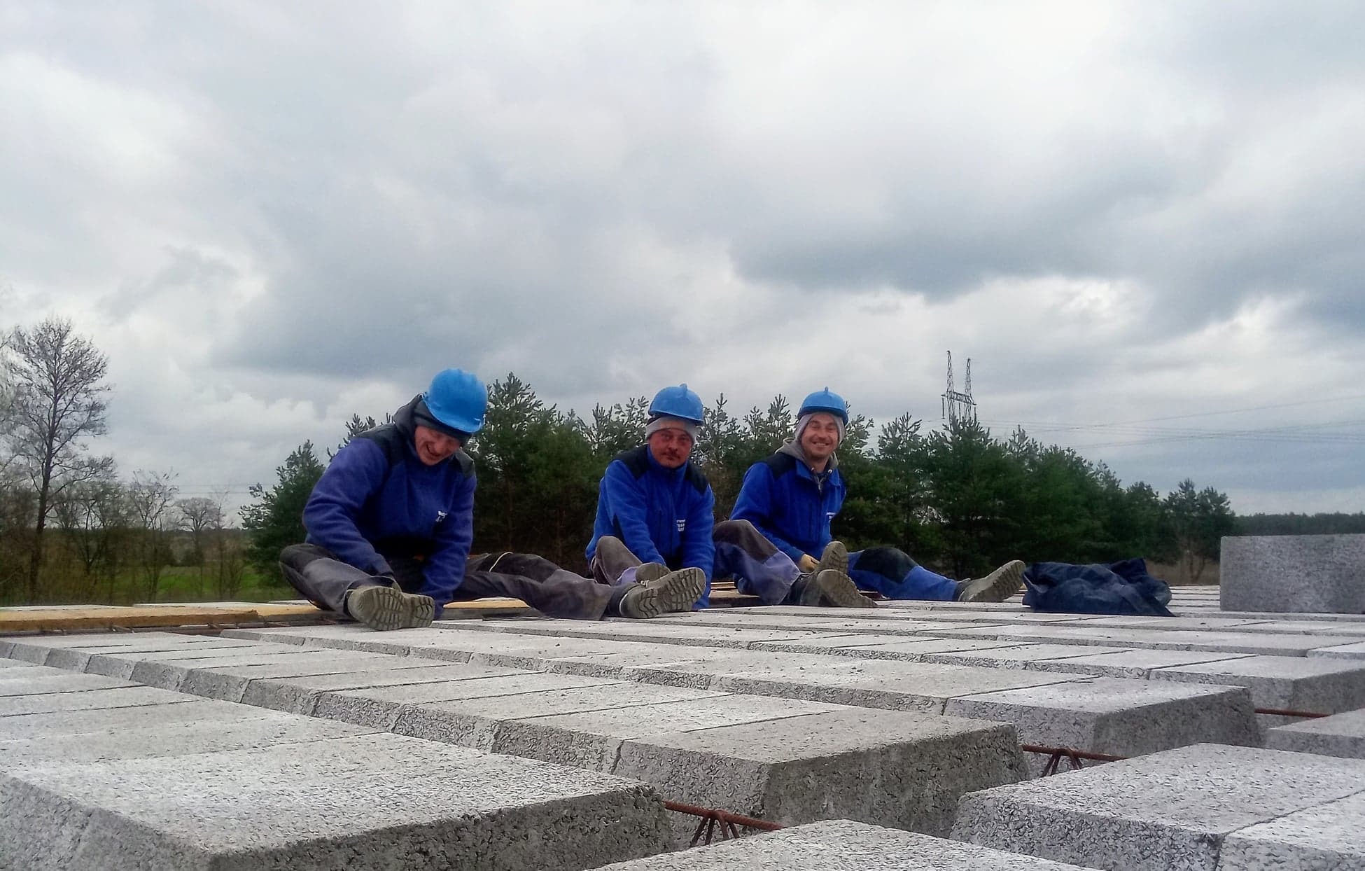 Trzech pracowników w niebieskich uniformach i kaskach siedzi na stropie z szarej kostki, w tle las i pochmurne niebo.