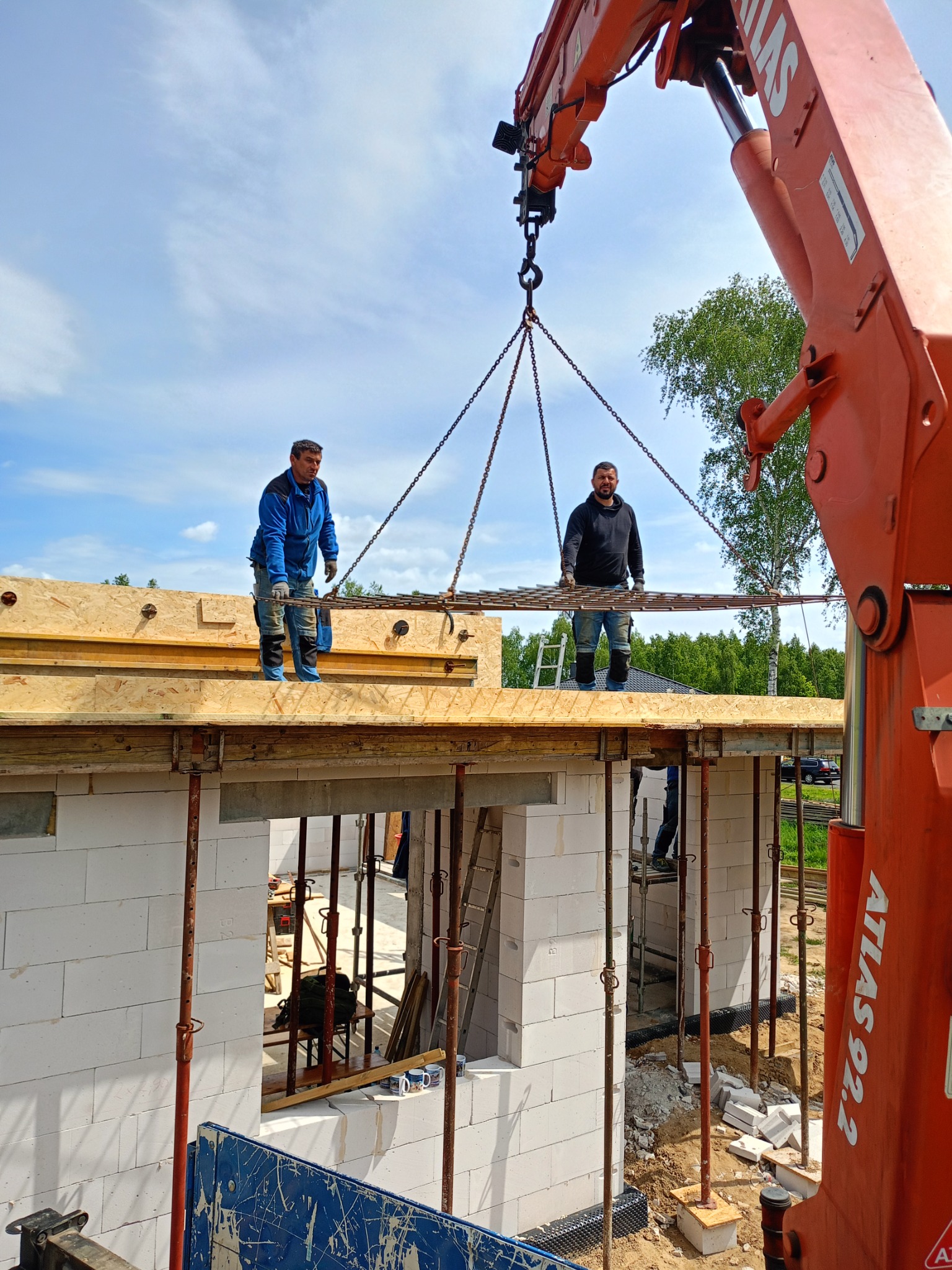 Budowa domu z betonu komórkowego: dwaj robotnicy na stropie, dźwig podnosi zbrojenie na tle błękitnego nieba z chmurami.