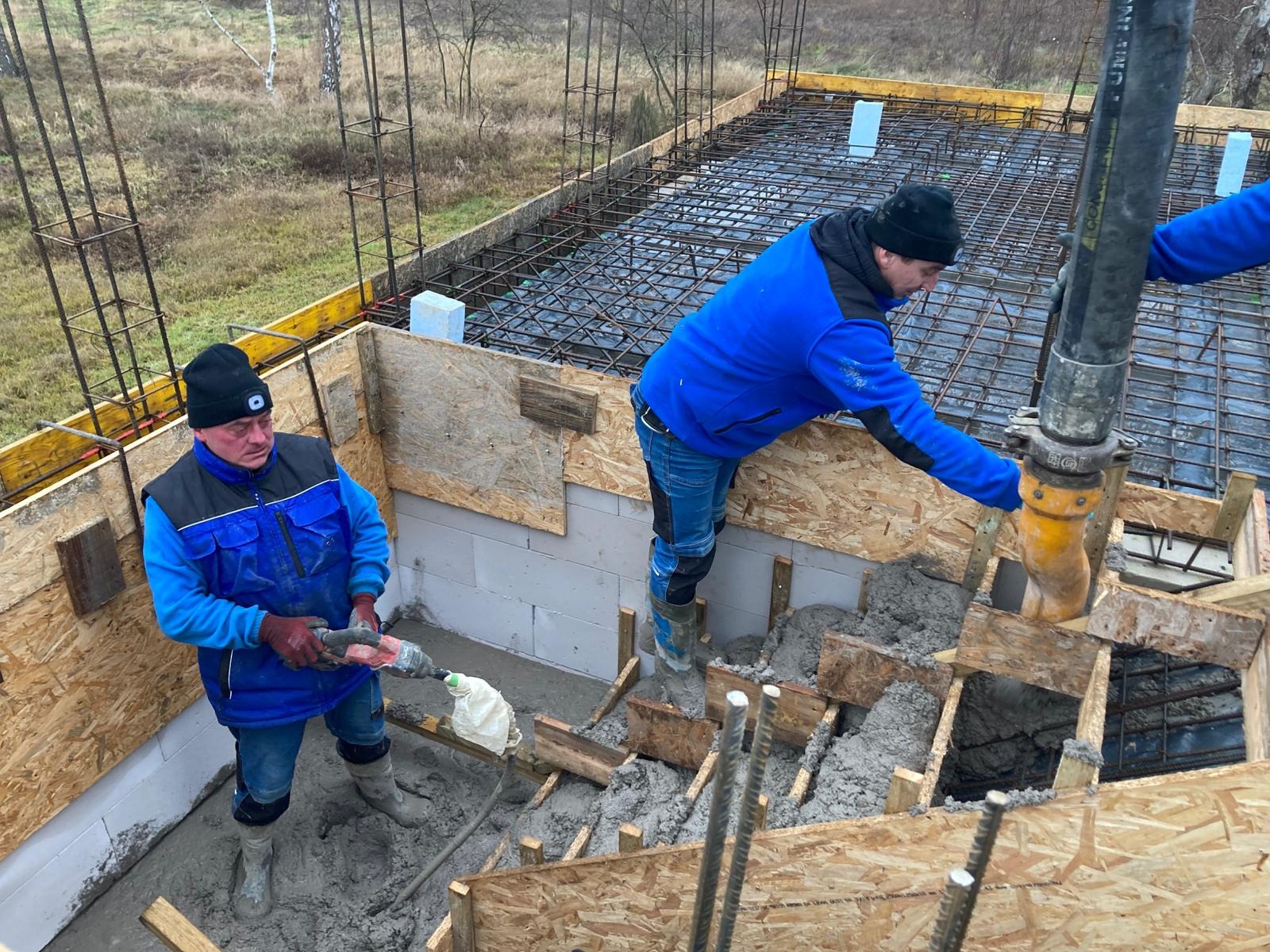 Wylewanie fundamentów domu z betonu. Dwóch robotników w trakcie pracy, jeden z wibratorem, drugi z pompą do betonu. Widoczne szalunki i zbrojenia.