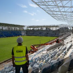 Pracownik w kamizelce odblaskowej i kasku, z napisem 'IZOLACJE PLUIMERS', obserwuje prace na budowie stadionu, gdzie widać podnośnik koszowy i przygotowane do montażu krzesełka pod folią.