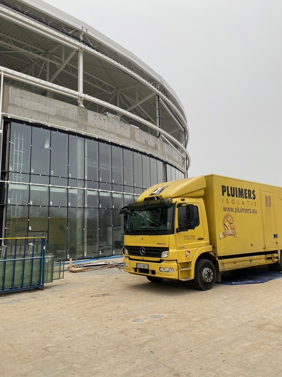 Żółta ciężarówka firmy izolacyjnej na tle budynku w trakcie budowy z dużą szklaną fasadą i metalową konstrukcją dachu.