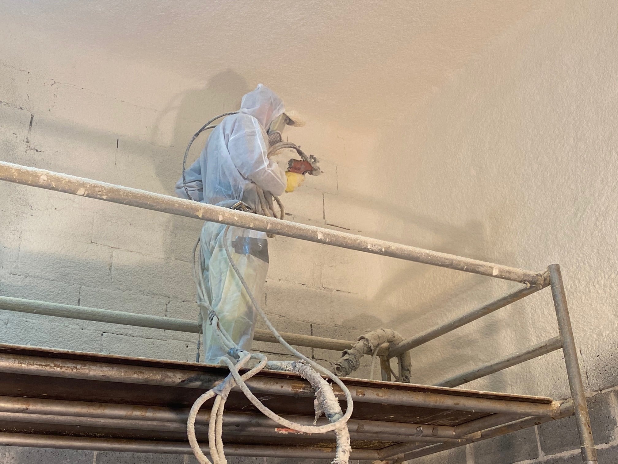 Pracownik w kombinezonie ochronnym na rusztowaniu aplikuje natryskowo izolację na ścianie z cegieł, widoczne przewody i narzędzie natryskowe.