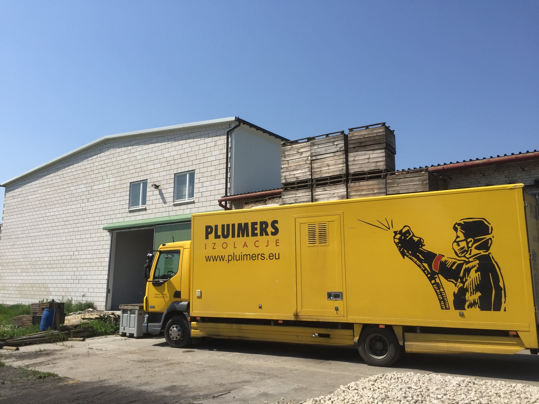 Żółta ciężarówka firmy z logo przedstawiającym osobę w masce ochronnej i kombinezonie, spryskującą coś pistoletem natryskowym, zaparkowana przed halą magazynową z drewnianymi skrzyniami na dachu