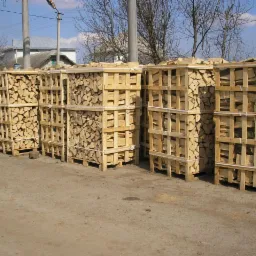 Rząd drewnianych skrzyń wypełnionych pociętym drewnem kominkowym, ustawionych na zewnątrz w słoneczny dzień. W tle budynki mieszkalne i drzewa bez liści.