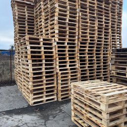Palety Malicki - Stos drewnianych palet ułożonych na zewnątrz, widoczne ślady użytkowania i naturalna faktura drewna. Tło stanowi szare niebo i fragment ogrodzenia.