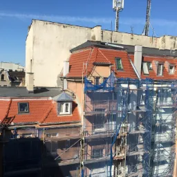 Budynek w rusztowaniach z niebieską siatką, widoczne fragmenty czerwonego pokrycia na dachu i anteny na sąsiednim budynku.