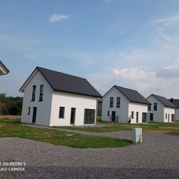 Firma ogólnobudowlana - Rząd nowoczesnych, białych domów z ciemnymi dachami, widoczny z perspektywy żwirowego podjazdu w słoneczny dzień, z ludźmi w tle i białym samochodem dostawczym po prawej.