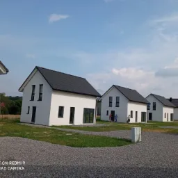 Rząd nowoczesnych, białych domów z ciemnymi dachami, widoczny z perspektywy żwirowego podjazdu w słoneczny dzień, z ludźmi w tle i białym samochodem dostawczym po prawej.