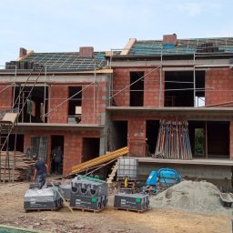 Firma ogólnobudowlana - Budowa bliźniaka z czerwonej cegły: widoczne rusztowania, częściowo położona dachówka, palety z dachówką BMI Braas na ziemi, betoniarka i robotnicy wewnątrz budynku.