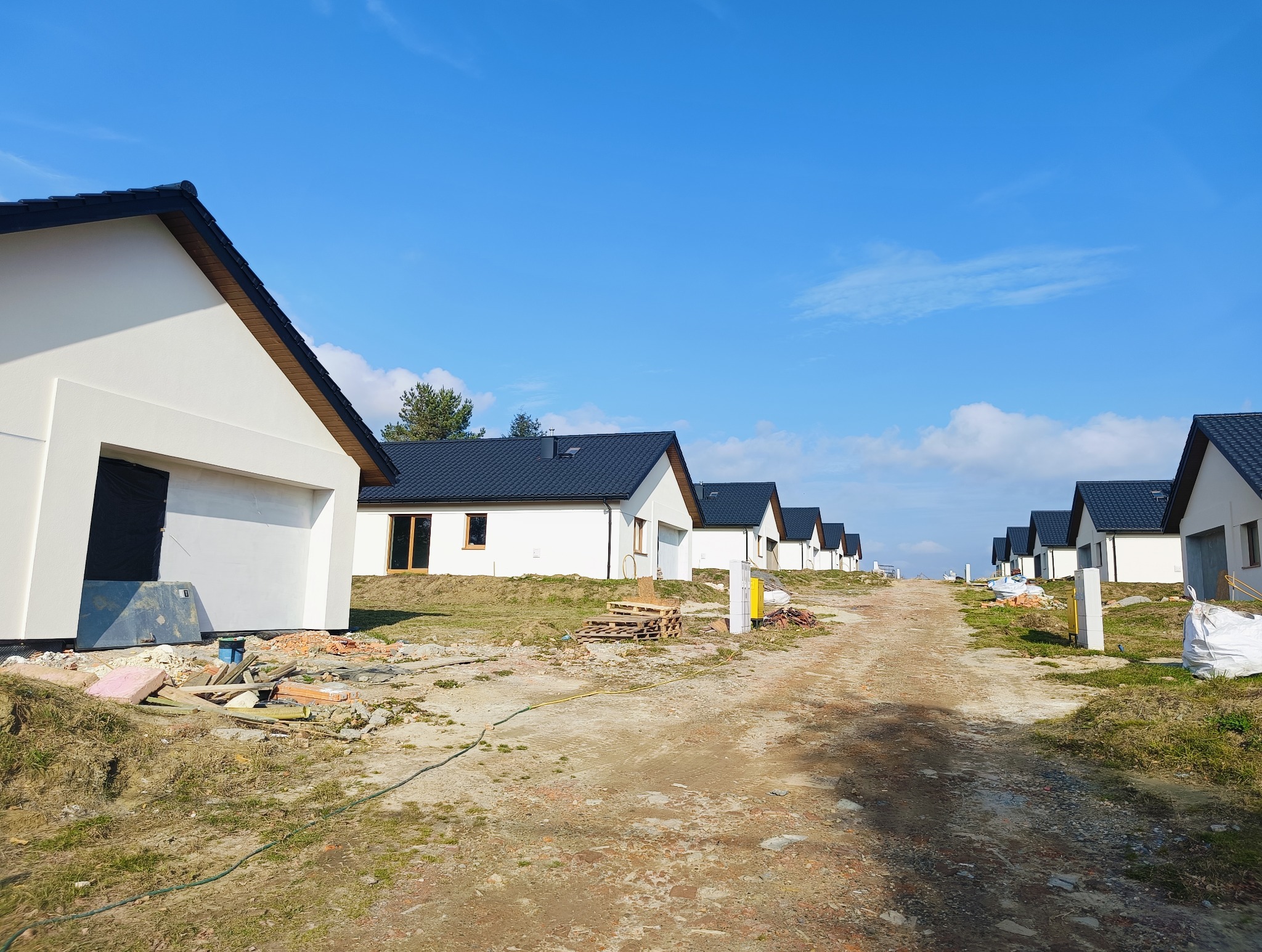 Szereg nowo wybudowanych, jednorodzinnych domów z białymi elewacjami i ciemnymi dachami, widok z drogi gruntowej w słoneczny dzień, teren budowy z materiałami i pozostałościami po budowie.