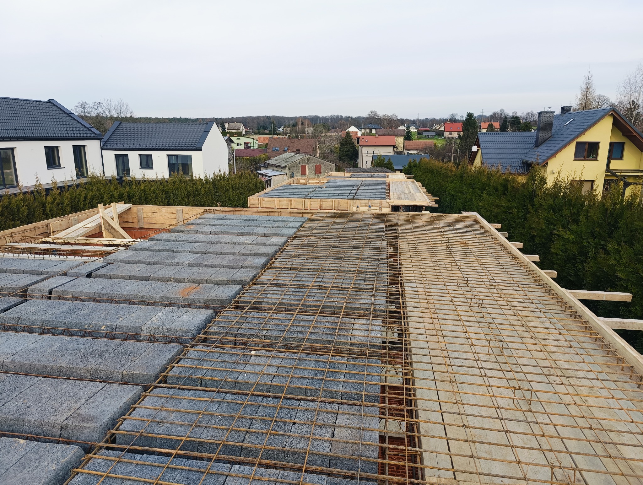 Wiązanie zbrojenia i układanie bloczków szalunkowych na budowie domu, widok z góry na konstrukcję stropu i otaczające budynki mieszkalne.