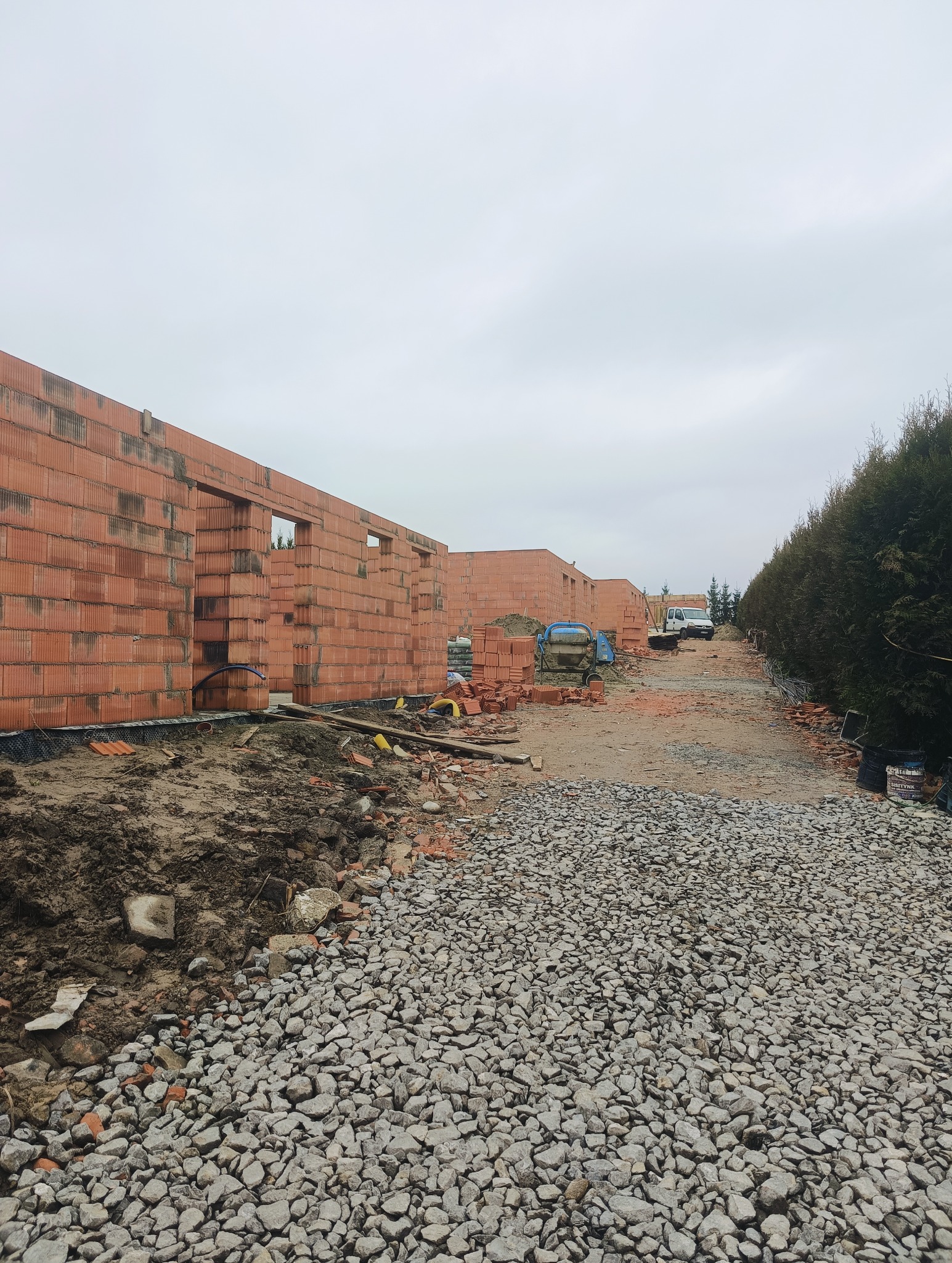 Wznoszenie ścian parterowego domu z czerwonej cegły ceramicznej, widoczne otwory okienne i drzwiowe, na pierwszym planie gruz i kamienie, w tle betoniarka i samochód dostawczy.