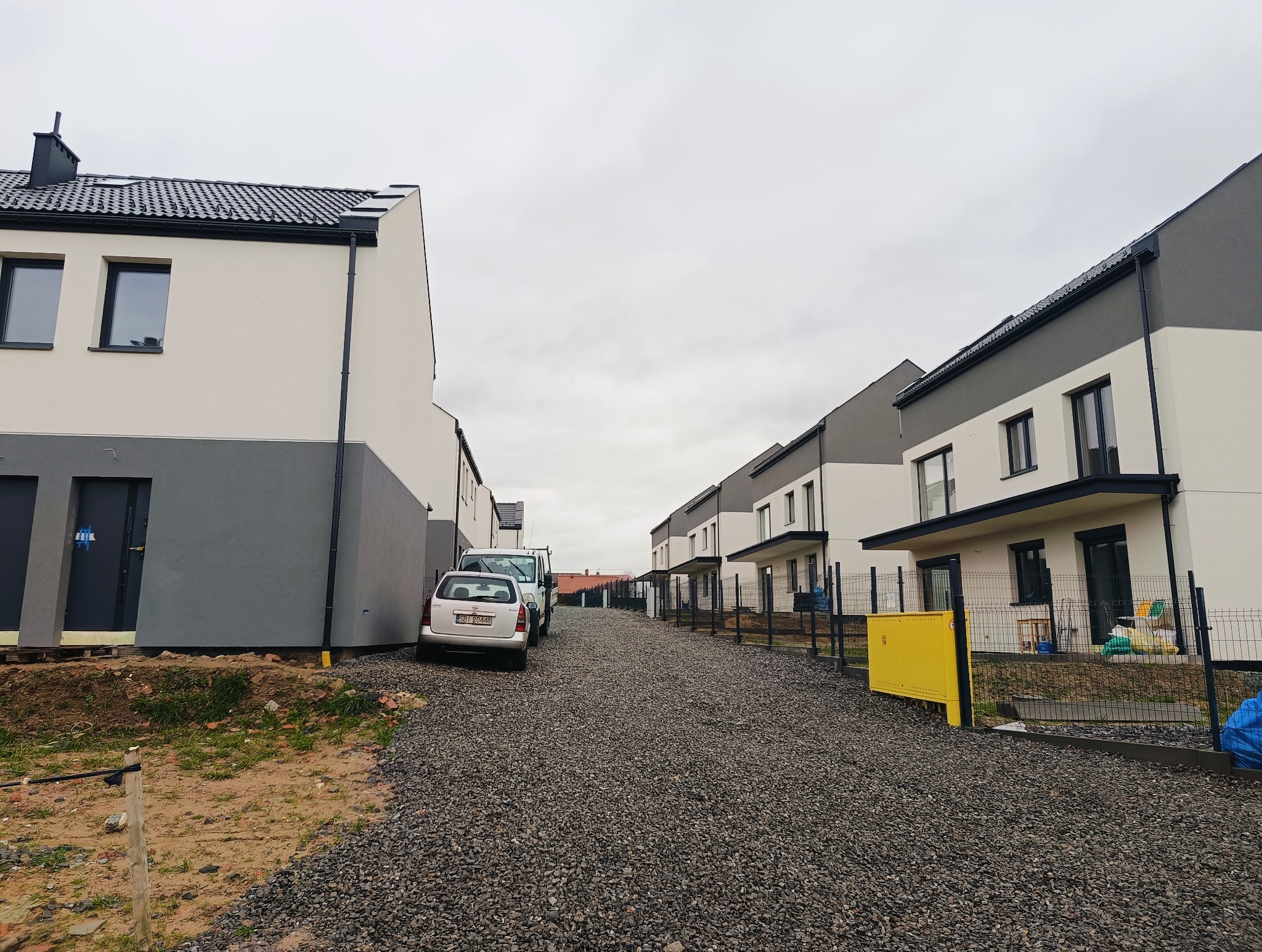 Nowe szeregowe domy murowane z dwukolorową elewacją (szary dół, biała góra) i czarną dachówką, widok z drogi szutrowej w pochmurny dzień.