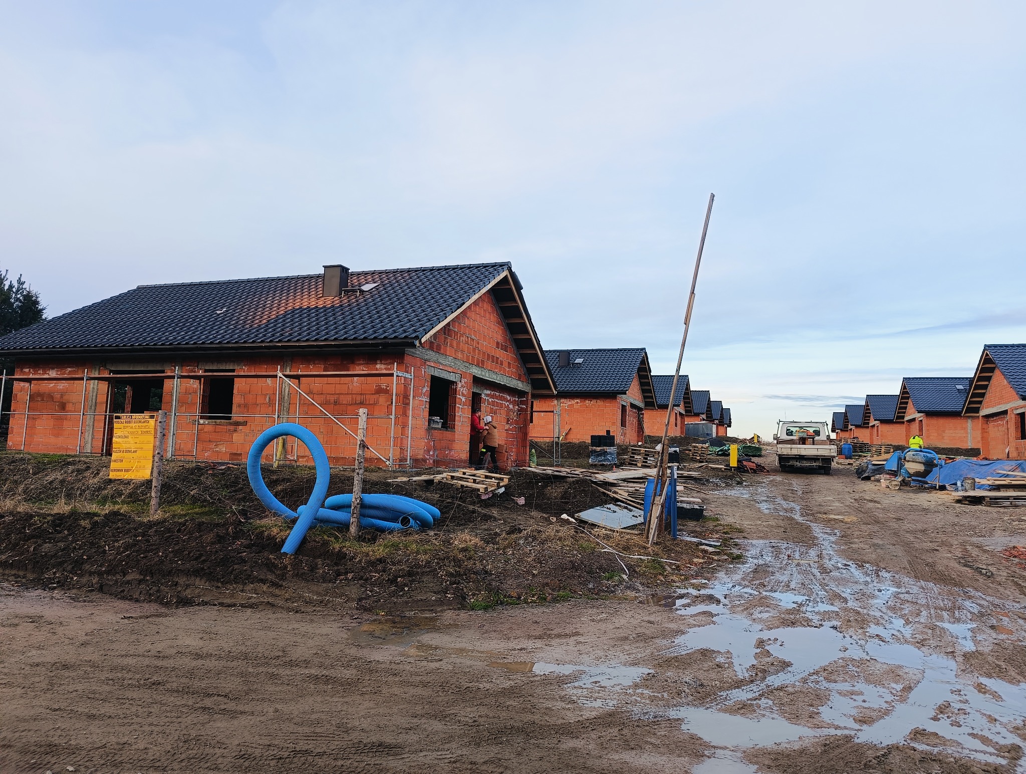 Budowa osiedla domów z czerwonej cegły, widoczne ślady błota po deszczu, niebieskie rury wentylacyjne zwinięte przy ogrodzeniu, w tle ciężarówka i betonowa mieszarka.