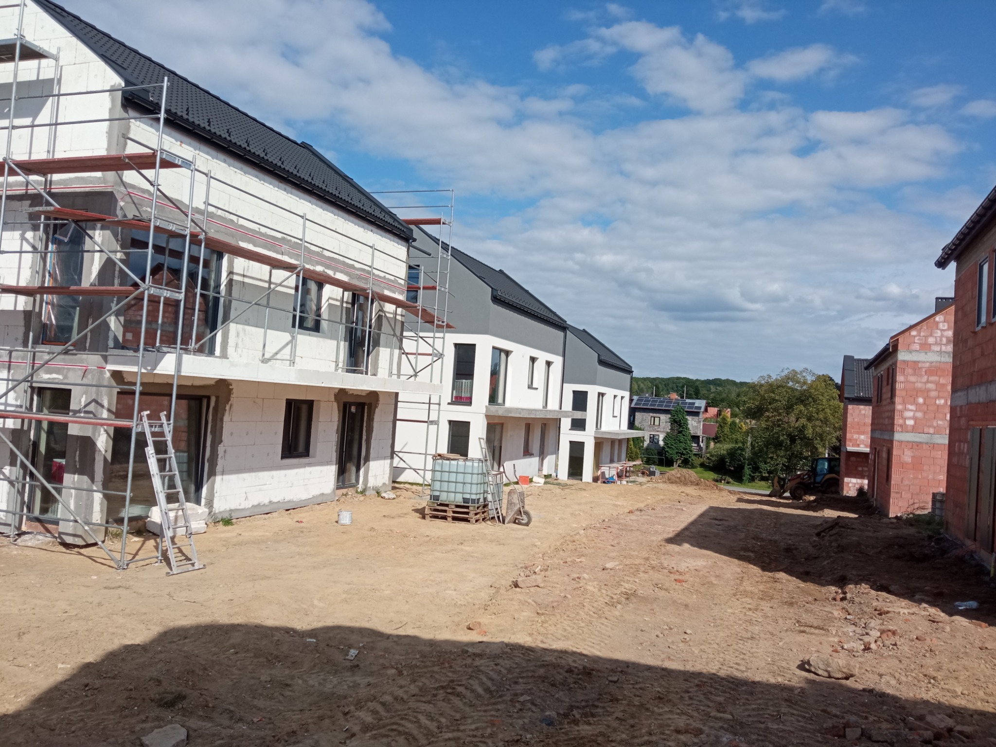 Wznoszenie szeregu domów w stanie surowym, z widocznymi rusztowaniami przy elewacjach, teren budowy z piaskiem i częściowo wykończony budynek z czerwonej cegły w tle.