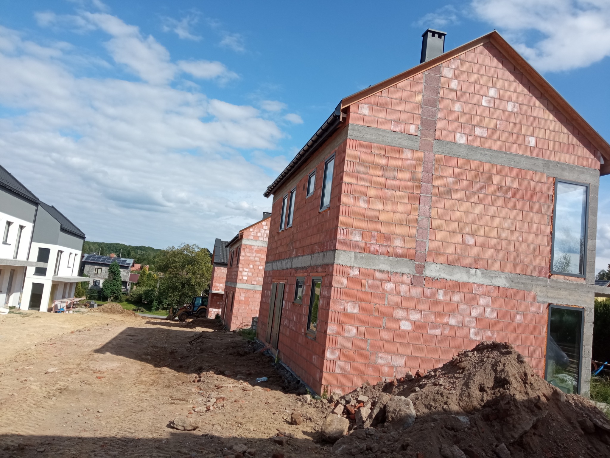 Wznoszenie ścian z czerwonej cegły na budowie szeregowców, widoczne okna, brak dachu, hałda ziemi na pierwszym planie, błękitne niebo w tle.