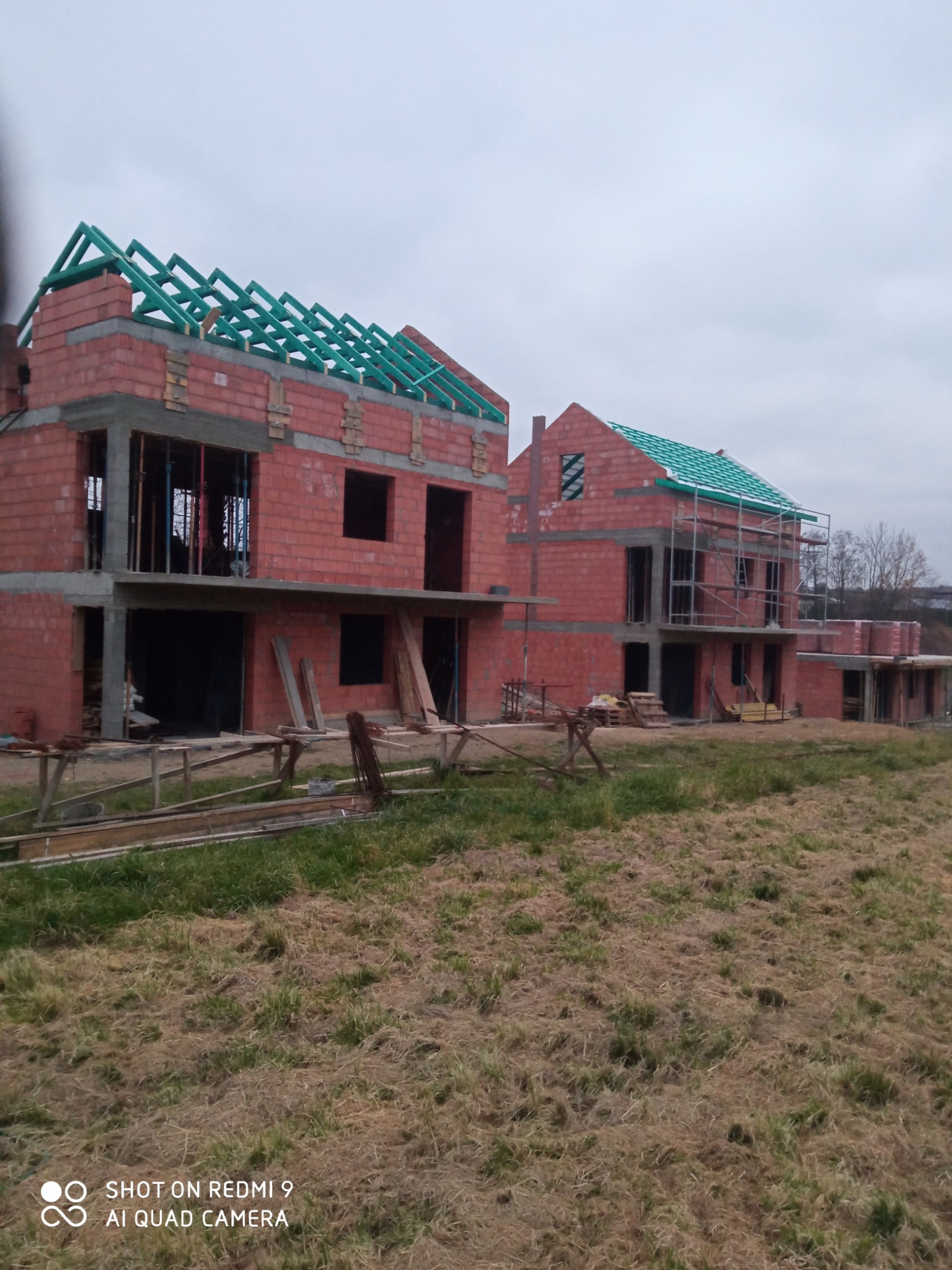 Dwa domy w trakcie budowy z czerwonej cegły z zamontowaną zieloną konstrukcją dachu, widoczne rusztowania i materiały budowlane.