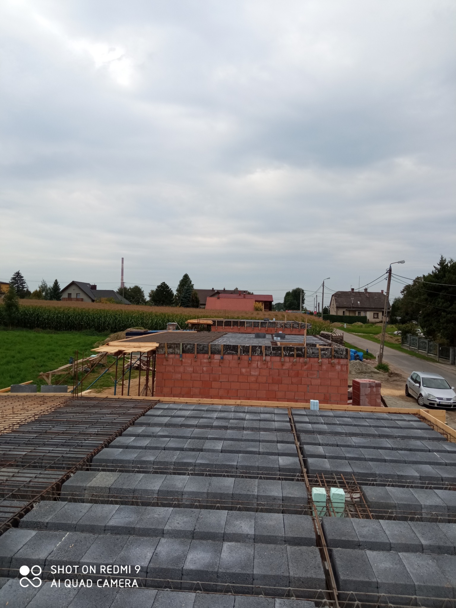 Wznoszenie ścian z czerwonej cegły na budowie domu, widok z góry na strop Teriva z szarymi bloczkami betonowymi, w tle krajobraz z polami i zabudowaniami, pochmurne niebo.