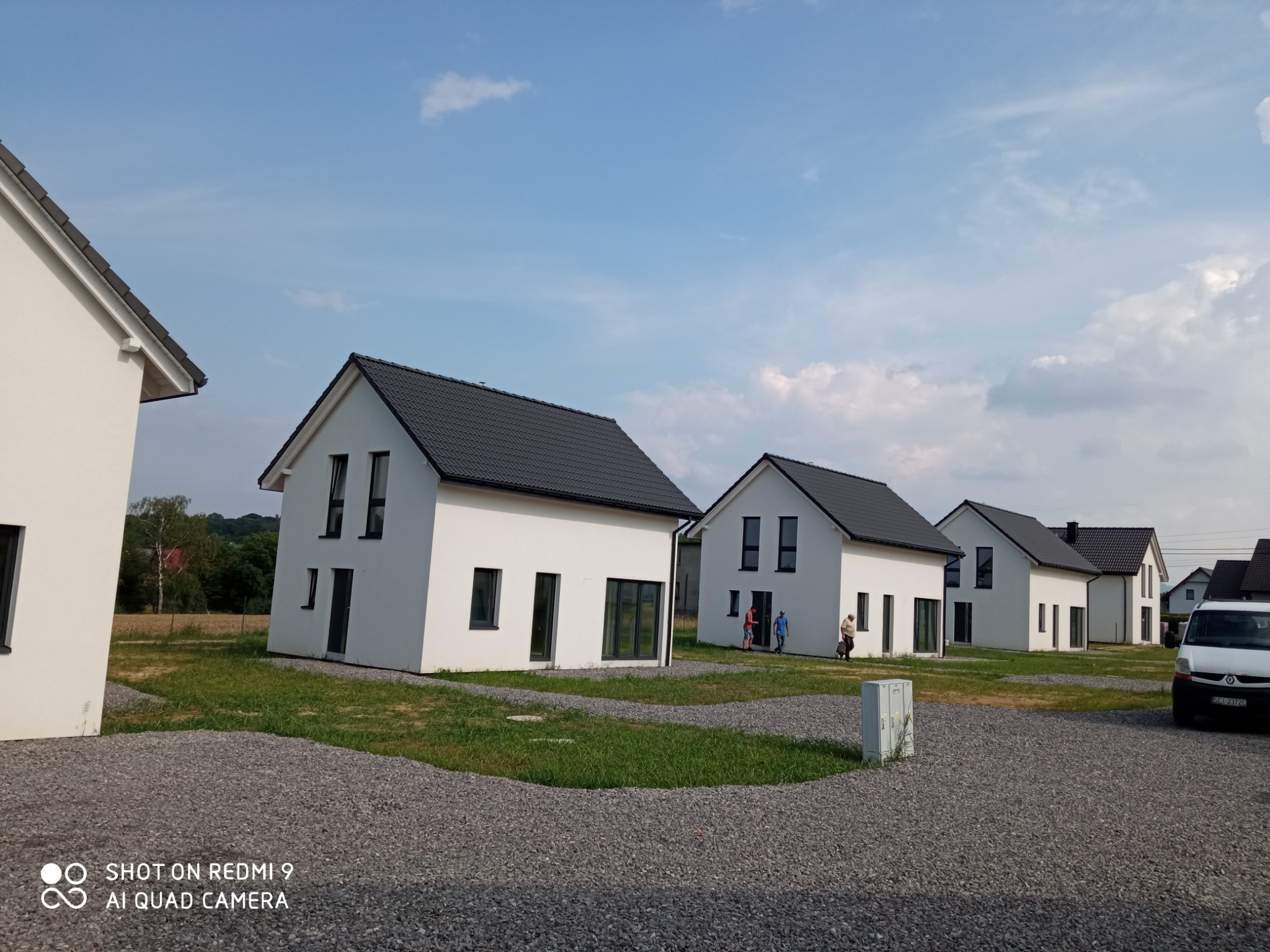 Rząd nowoczesnych, białych domów z ciemnymi dachami, widoczny z perspektywy żwirowego podjazdu w słoneczny dzień, z ludźmi w tle i białym samochodem dostawczym po prawej.