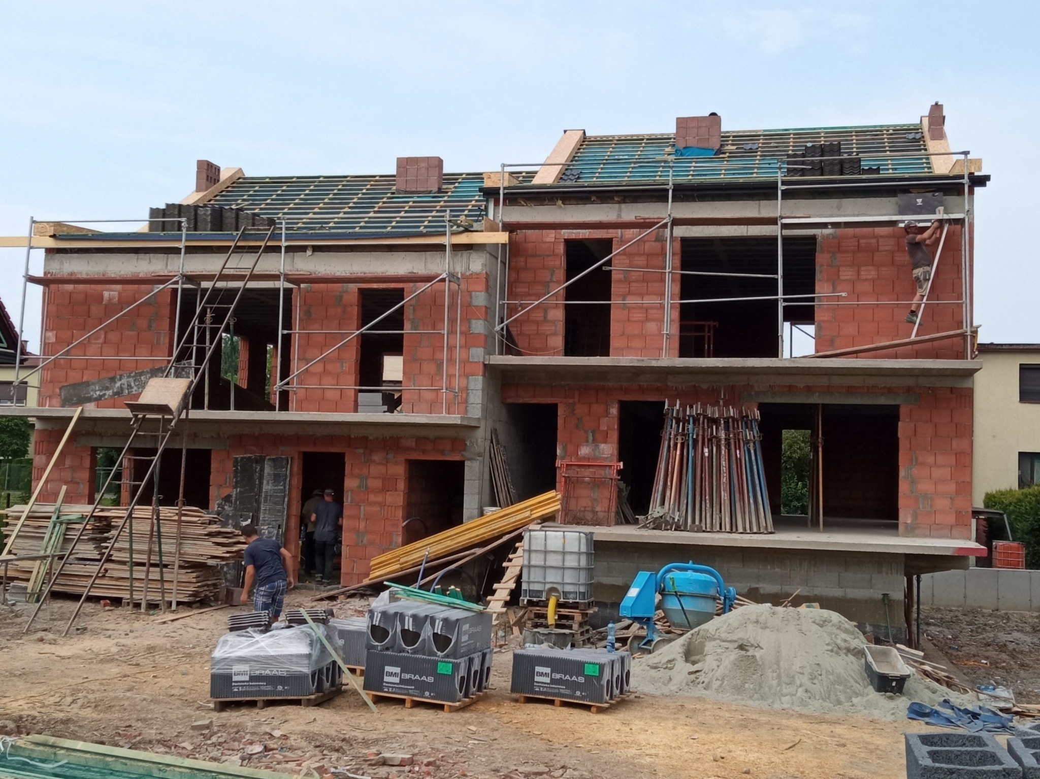 Budowa bliźniaka z czerwonej cegły: widoczne rusztowania, częściowo położona dachówka, palety z dachówką BMI Braas na ziemi, betoniarka i robotnicy wewnątrz budynku.