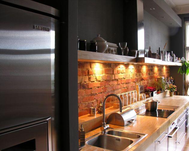 Kuchnia z blatem z betonu architektonicznego, ceglaną ścianą i oświetleniem punktowym pod półką, widoczny zlew dwukomorowy i sprzęt AGD ze stali nierdzewnej.