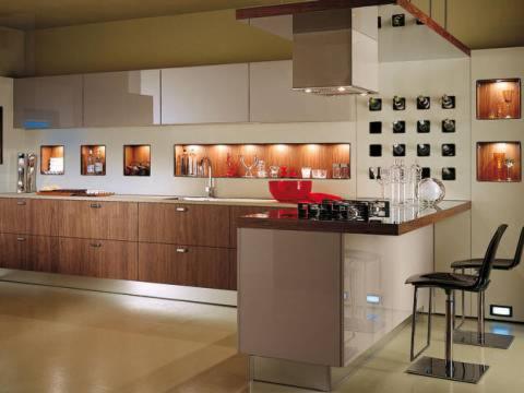 Nowoczesna kuchnia z drewnianymi frontami szafek dolnych i jasnymi górnymi, podświetlane wnęki z dekoracjami, wyspa z płytą grzewczą i barkiem oraz czarne hokery na metalowych nogach.