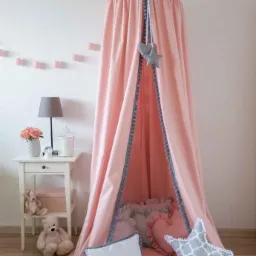 R&oacute;żowy baldachim z szarymi pomponami nad okrągłą matą i poduszkami w pokoju dziecięcym, obok biały stolik z lampką i dekoracjami.