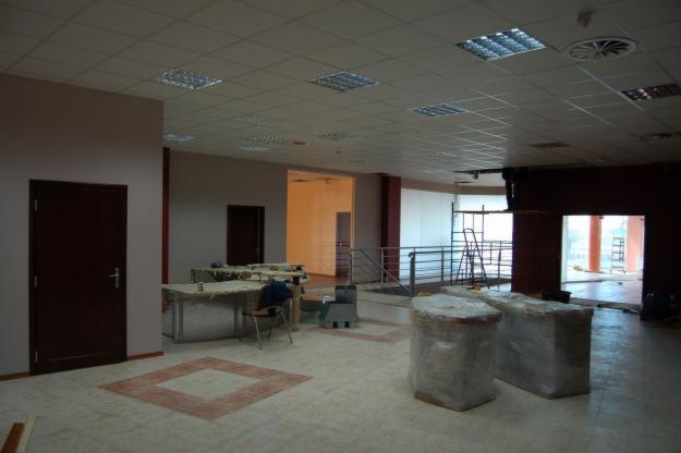 Wnętrze dużego pomieszczenia w trakcie prac wykończeniowych, z widocznymi elementami suchej zabudowy zabezpieczonymi folią, stolikami warsztatowymi i rozłożonymi narzędziami, podwieszanym sufitem...