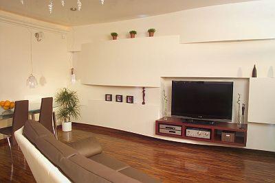 Nowoczesny salon z drewnianą podłogą, białą ścianą z wnęką na telewizor i półkami, widoczne elementy suchej zabudowy, stół z owocami i minimalistyczne lampy wiszące.