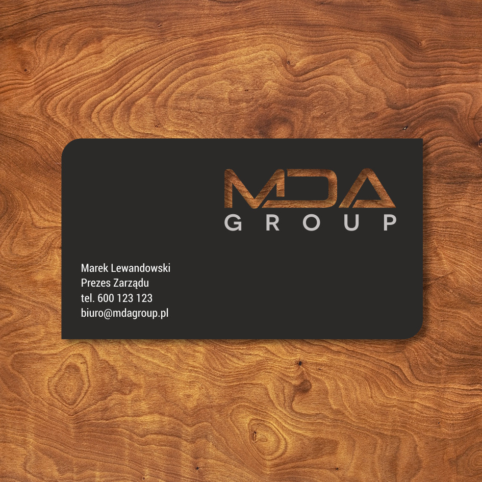 Elegancka, czarna wizytówka firmy MDA Group z drewnianym wypełnieniem liter logo, leżąca na tle z teksturą drewna.