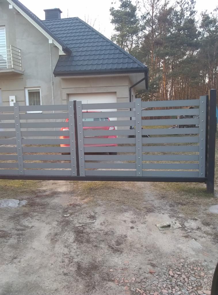 Nowoczesna, przesuwna brama w kolorze szarym z poziomymi panelami, zamontowana przed domem z szarą elewacją i ciemnym dachem, z widocznym fragmentem czerwonego samochodu w tle.