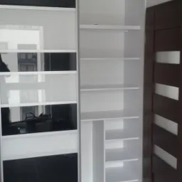 Biała szafa przesuwna z czarnymi i białymi panelami szklanymi, widoczne półki i system prowadnic, minimalistyczny design.