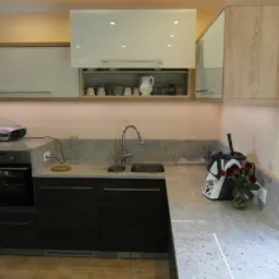 Nowoczesna kuchnia z ciemnymi frontami szafek, jasnym blatem granitowym, i górnymi szafkami z białymi frontami oraz drewnopodobnym korpusem. Na blacie stoi robot kuchenny i wazon z czerwonymi różami.