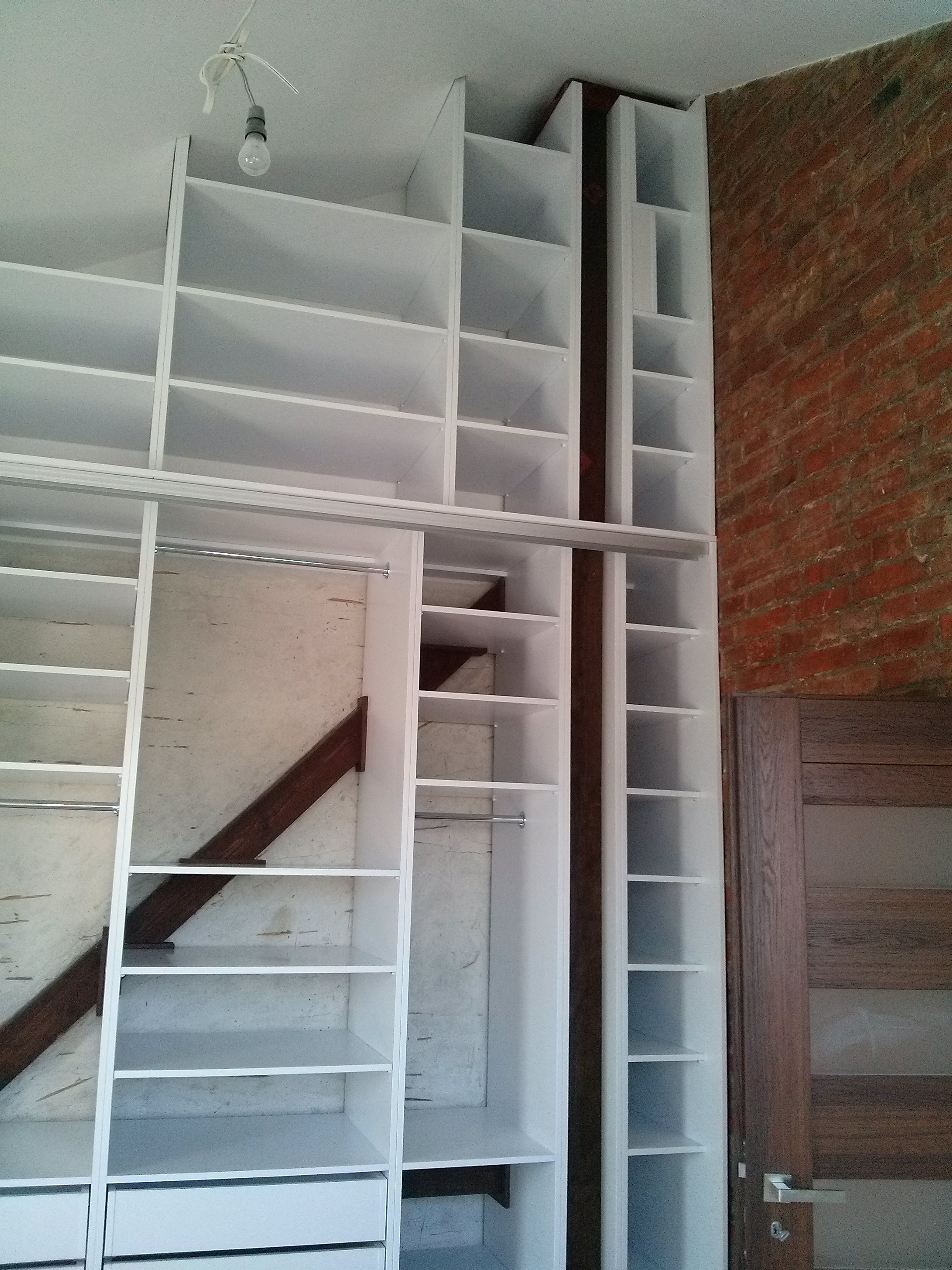 Biała szafa wnękowa na wymiar z drążkiem i półkami, dopasowana do skosu poddasza, z widoczną ceglaną ścianą i drzwiami w tle.