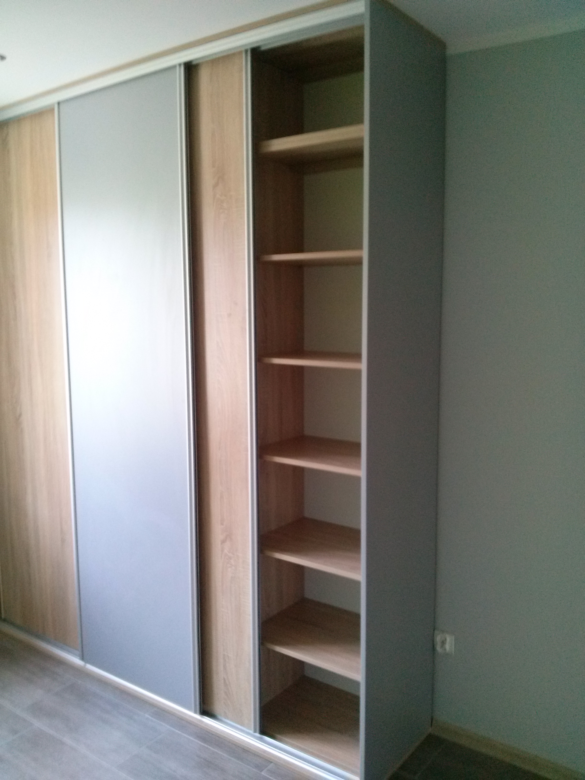 Szafa wnękowa z przesuwnymi drzwiami, wykończona w odcieniach drewna i szarości, z otwartym regałem z półkami po prawej stronie.