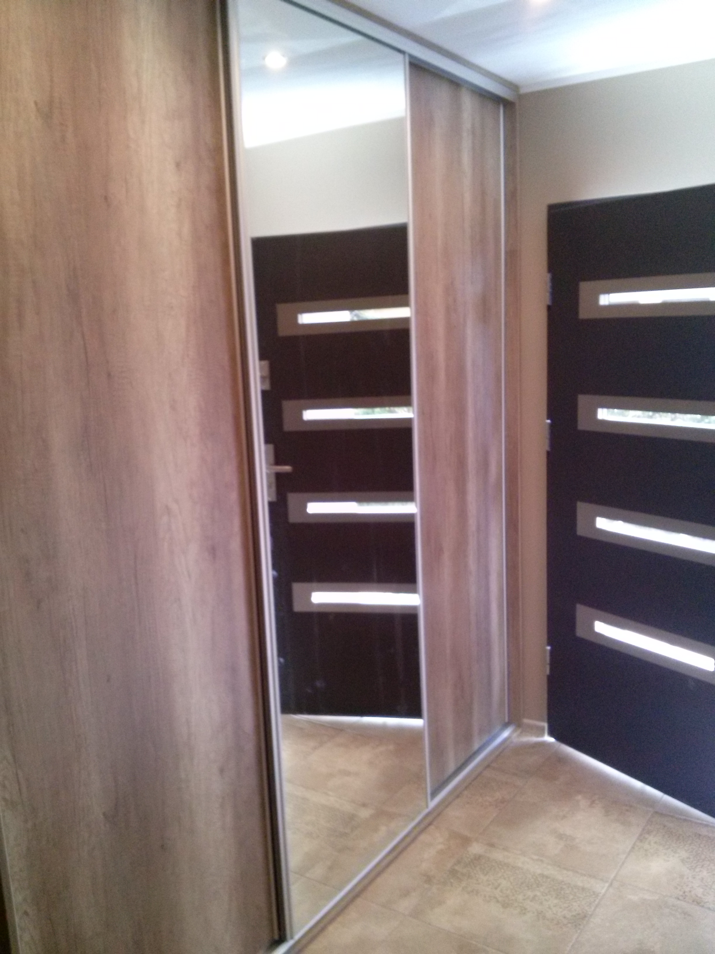 Szafa wnękowa z lustrzanymi drzwiami przesuwnymi, wykończona okleiną drewnopodobną. W lustrze odbija się drzwi z poziomymi przeszkleniami.