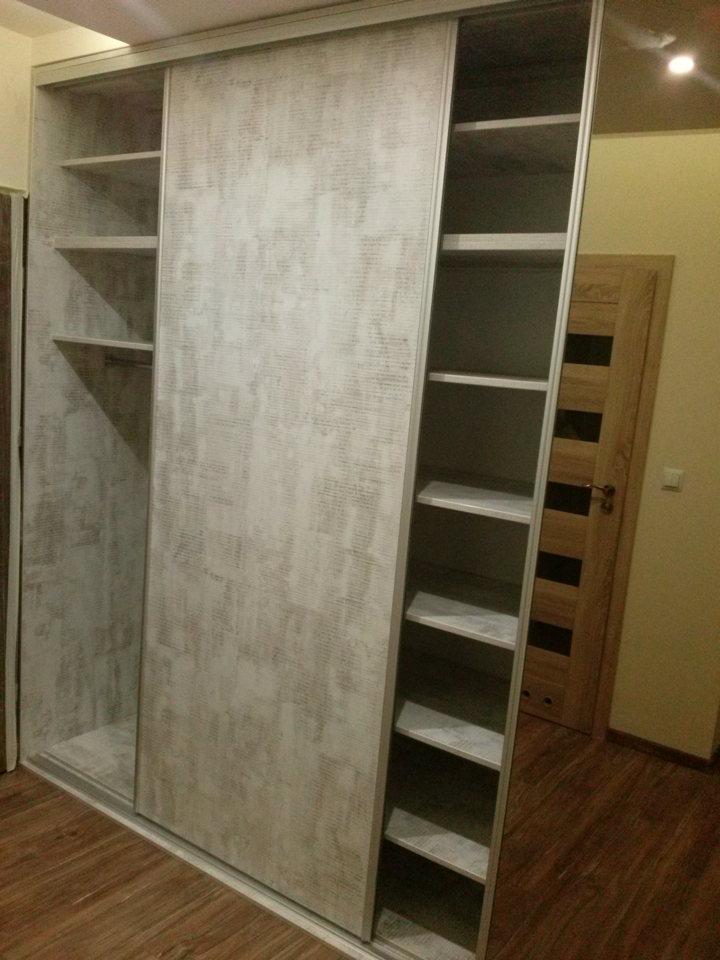 Szafa wnękowa z przesuwnymi drzwiami w odcieniach bieli i szarości, z widocznymi półkami i drążkiem na ubrania. Widoczne drewniane panele podłogowe.