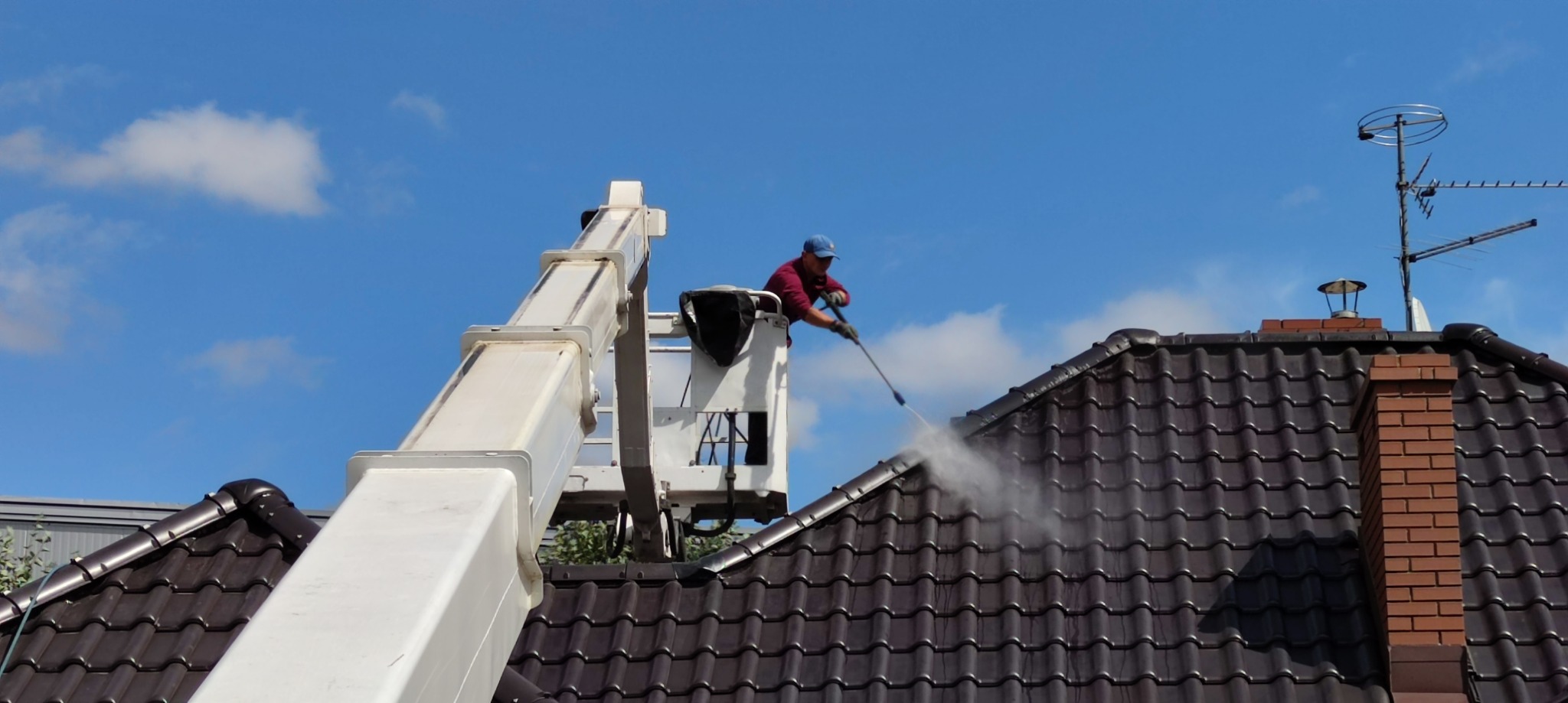 Pracownik w podnośniku koszowym czyści dach z brązowej dachówki za pomocą myjki ciśnieniowej, widoczny komin z cegły i antena telewizyjna na tle błękitnego nieba.