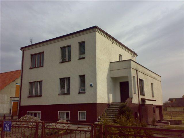 Dwupiętrowy budynek z białej cegły z ciemnobrązową podmurówką i ramami okiennymi, widoczny zza brązowego metalowego ogrodzenia. Na pierwszym planie stos gruzu.