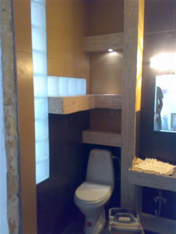 Remont łazienki w Inowrocławiu: Widok na toaletę, półki z surowego drewna z oświetleniem punktowym, ścianę z luksferów i fragment lustra z odbiciem osoby.