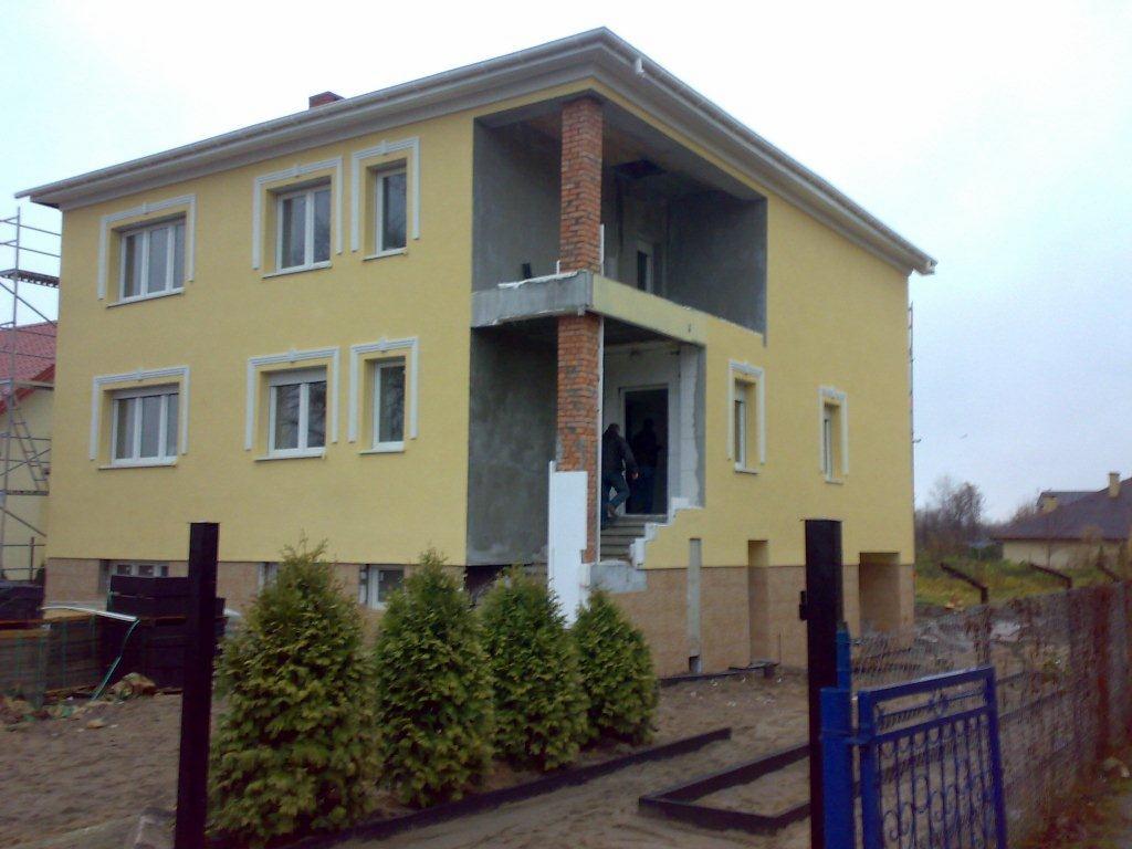 Żółty dom dwupiętrowy w trakcie ocieplania, z widocznym rusztowaniem i elementami izolacji na ścianach, z trzema niewielkimi drzewkami iglastymi na pierwszym planie.