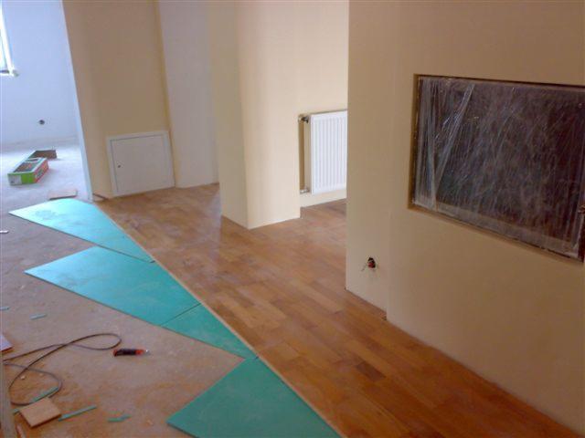 Układanie parkietu: widok na fragment podłogi z ułożonymi deskami parkietowymi, częściowo przykrytymi zielonym podkładem, narzędzie do cięcia leży obok, okno zabezpieczone folią malarską.