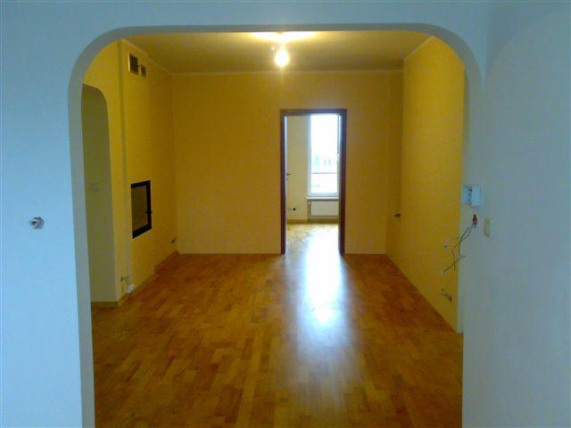 Pusty pokój z parkietem, żółtymi ścianami i łukowym przejściem, widok na kolejne pomieszczenie z oknem.