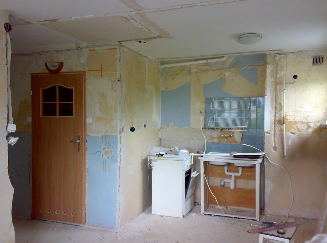 Wnętrze kuchni w trakcie remontu: zerwane tapety, odsłonięte instalacje elektryczne, częściowo zdemontowane szafki kuchenne i stara kuchenka. Widoczne ślady po starej instalacji elektrycznej...