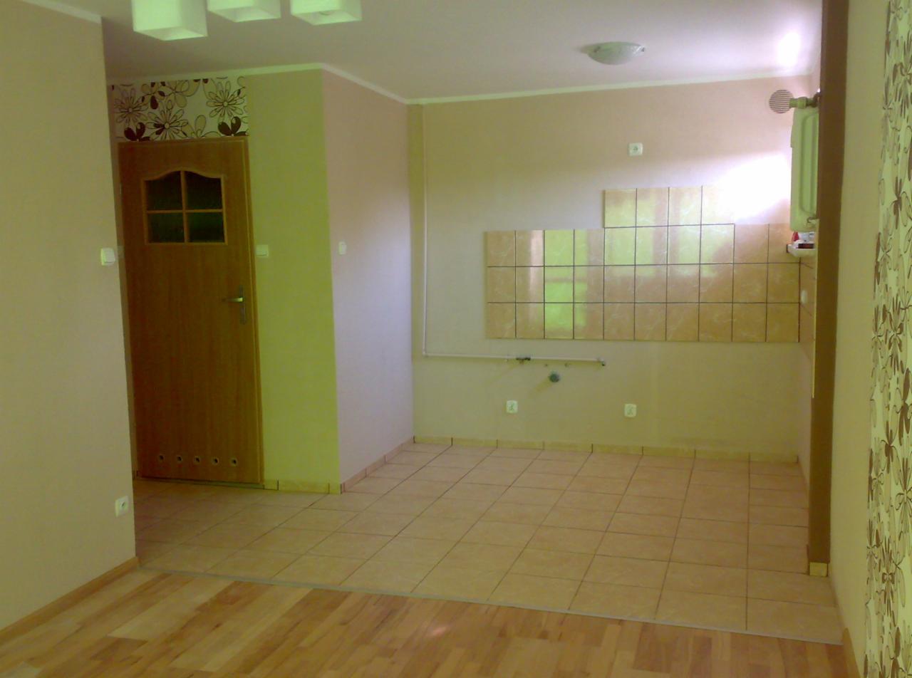 Wnętrze pokoju w trakcie remontu: beżowe płytki na ścianie, brązowe płytki na podłodze, drewniane drzwi z szybkami, ściany w odcieniach beżu i zieleni, widoczna instalacja wodna.