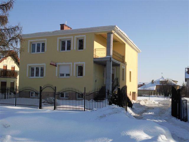 Dwupiętrowy budynek z żółtą elewacją i balkonem, otoczony czarnym metalowym ogrodzeniem, w zimowej scenerii ze śniegiem.