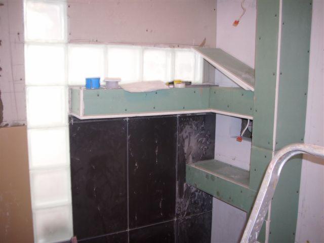 Łazienka w trakcie remontu: konstrukcja z płyty gipsowo-kartonowej obudowująca instalacje, czarne płytki na ścianie, luksfery w oknie, widoczne przewody elektryczne.