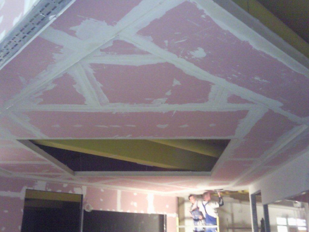 Montaż sufitu podwieszanego z płyt kartonowo-gipsowych z widocznym otworem i pracownikami na drabinie podczas szpachlowania łączeń.