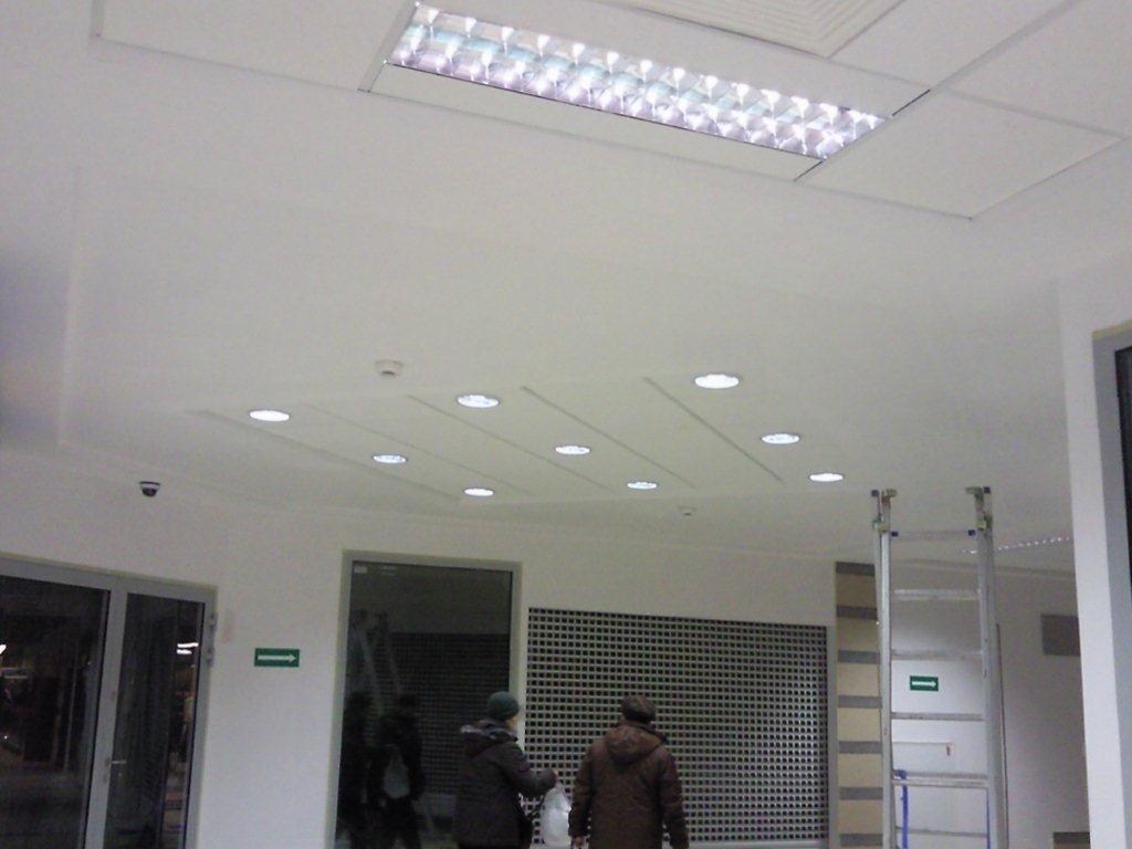 Wykończony sufit podwieszany z płyt kartonowo-gipsowych z wbudowanym oświetleniem punktowym i kasetonami, widoczne dwie osoby i drabina w tle.